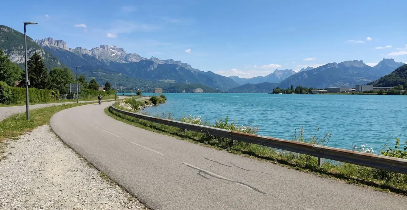 Dedicated cycling path curving alongside Lake Annecy's turquoise waters with distant Alpine peaks
